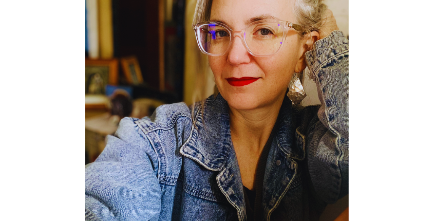 Headshot of Beth Watts Nelson, a woman with silver hair and clear-framed glasses, wearing a denim jacket and red lipstick.