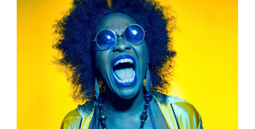 Headshot of performing artist BodaciousThang, a woman with a large natural afro and round glasses, mouth open wide, wearing blue lipstick and bold beaded necklaces against a bright yellow background.