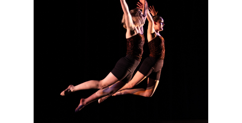 Two City in Motion dancers leaping in unison against a black background, arms raised and legs extended, wearing matching dark costumes. Photo credit Philip Koenig.