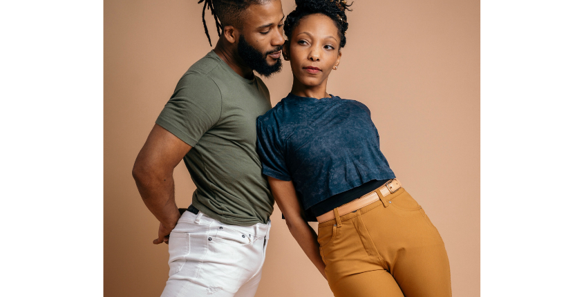 Two DynamitExperience dancers, a man and a woman, pose closely together against a warm beige background, both wearing casual streetwear. Photo Credit Becca Vision.