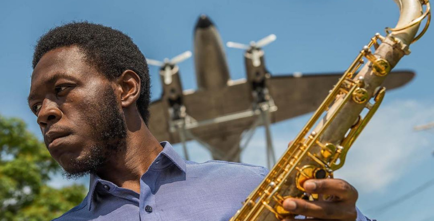 Headshot of Ernest Melton, a man holding a saxophone outdoors with a vintage aircraft visible in the background against a blue sky.