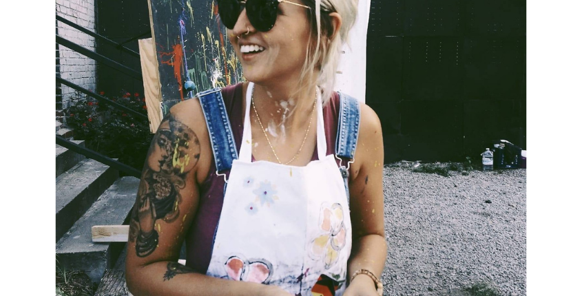 A smiling woman with short blonde hair and sunglasses sits outdoors in a paint-splattered apron, with a colorful canvas visible behind her.