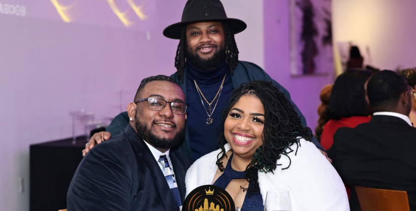 Three people smile together at an event, posed in front of a purple-lit backdrop bearing the Echoes of the City logo.