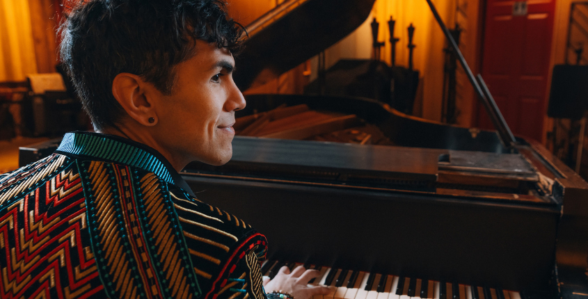 Ryan Marquez plays a grand piano in profile, wearing a bold patterned blazer in a warmly lit studio space.