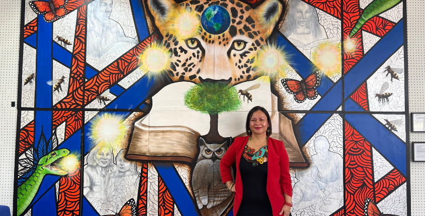 Artist Socorro Reyes Ramirez stands in front of her large mural featuring a jaguar, an open book, butterflies, and other vivid imagery, wearing a red blazer.