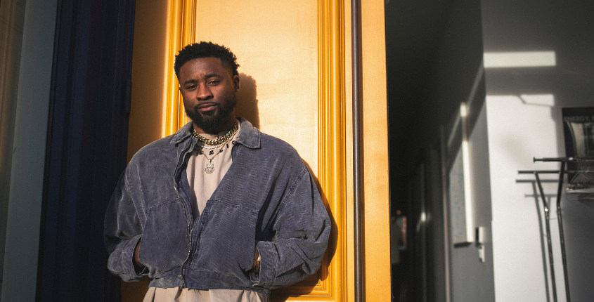 A man stands in a doorway framed by a bold yellow door, wearing a blue corduroy jacket and layered gold chains.
